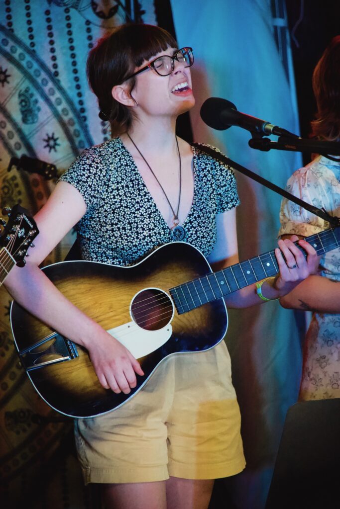 Lauren, with her brown hair in a bun, straight-across bangs, and black glasses, wearing yellow shorts and a flower-patterned black and white top, sings into a mic with her eyes closed. She is playing her vintage acoustic guitar, which is black along the edges with an orange-ish brown sunburst in the middle and a cream-colored pickguard.