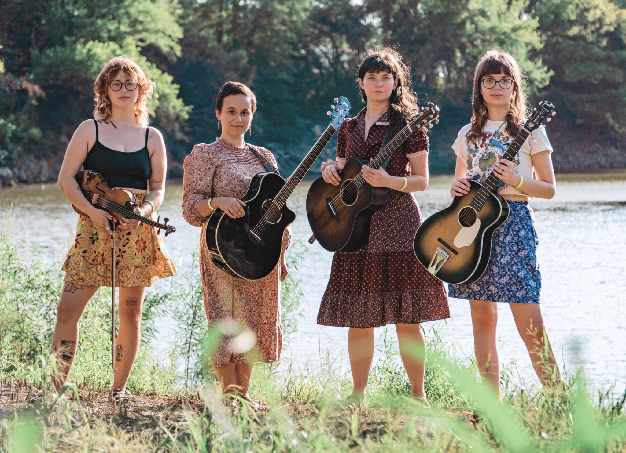 Four friends stand side by side with grass in the foreground and water in the background. From left to right: Gray, a person in a yellow, floral-patterned skirt and black tank top with black glasses and tattoos, holds a fiddle; Siena, a person with short, dark brown hair, pointy dangling earrings, and a mauve patterned long dress, holds a black guitar; Emily, a woman with long brown hair with straight-across bangs and a patterned burgundy dress holds a dark brown guitar; and Lauren, a woman with mid-length brown hair with straight-across bangs and black glasses, wearing an indigo skort and beige cropped t-shirt, holds a vintage sunburst guitar.