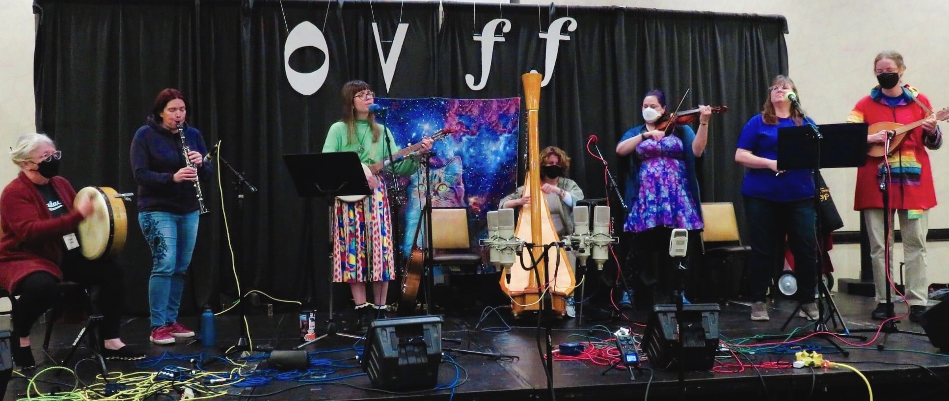 Seven women play as a band on stage; from the left, a woman with a red cardigan and white hair plays an Irish drum, an auburn-haired woman wearing jeans and a dark purple hoodie plays clarinet, a woman wearing a green long-sleeved shirt and multicolored skirt plays banjo and sings, a woman with short and wavy brown hair plays a big pedal harp, a woman with purple and blue hair in a matching dress plays violin, a brown-haired woman in a blue t-shirt and jeans looks up into the distance, and a tall woman in a red jacket with rainbow sleeves plays mandolin.