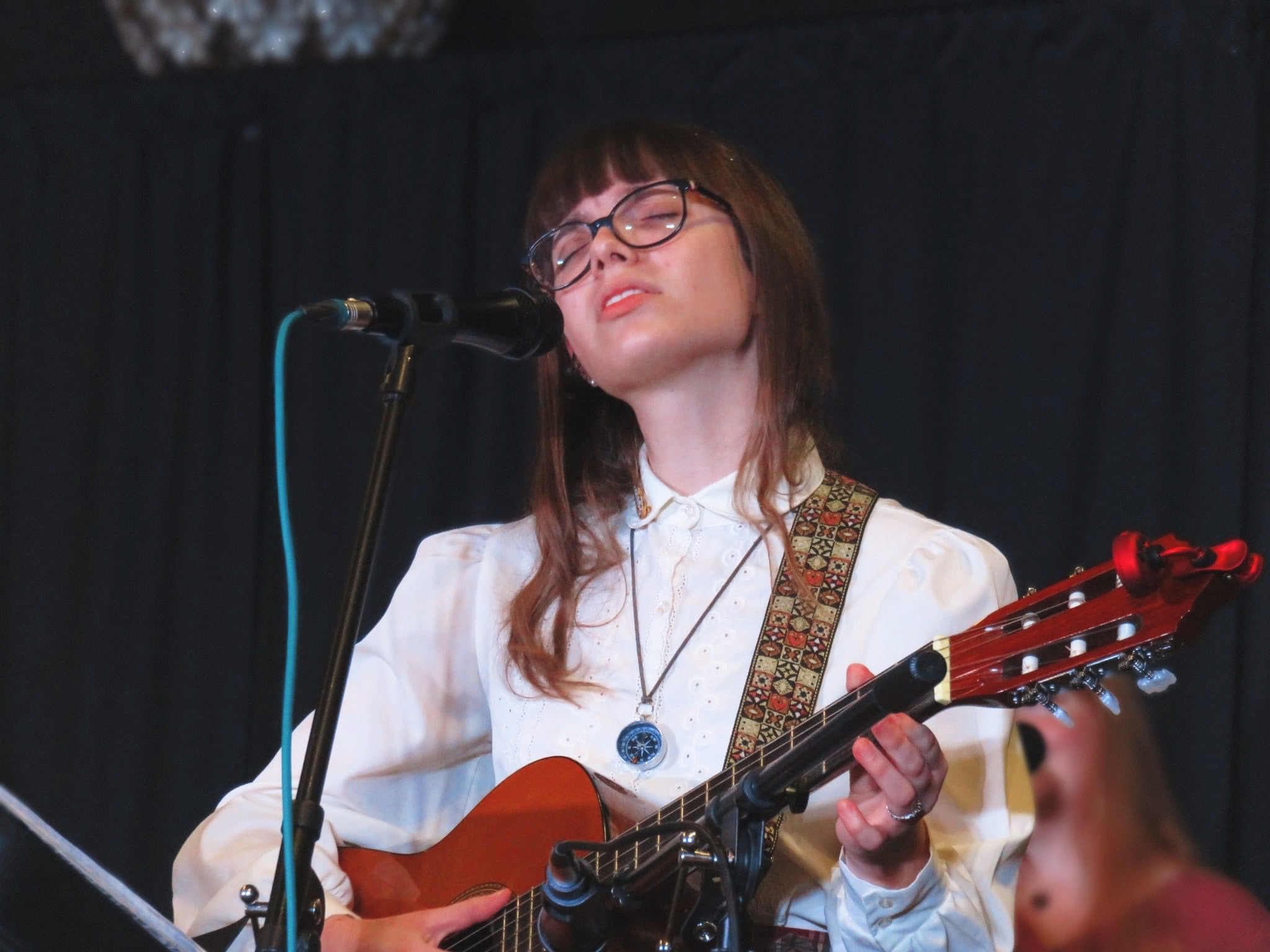 Lauren sings into a microphone with her eyes closed. She is playing a guitar and is wearing an ivory buttoned-up blouse, along with her compass necklace.